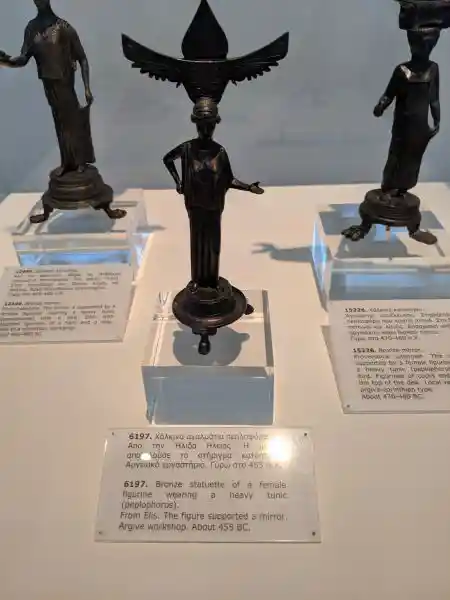 Ancient Greek bronze statuette of female figure wearing heavy tunic and supporting mirror with winged ornament on head